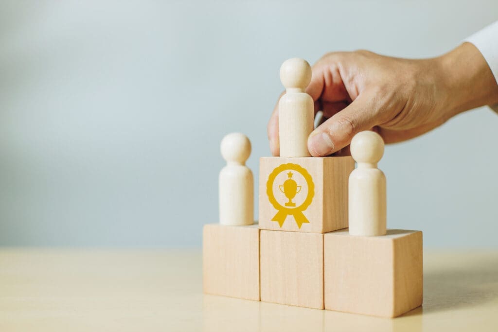 A hand places a wooden figure on a podium made of wooden blocks. One block has a yellow trophy symbol, indicating first place. Two more figures stand on lower blocks, representing second and third places.