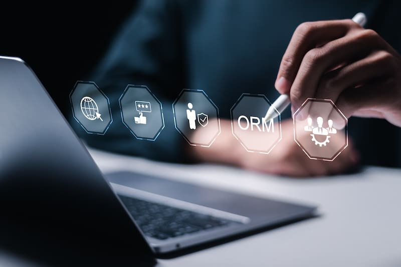 A person using a stylus interacts with a series of icons projected above a laptop keyboard. The icons depict a globe, a star rating, a figure with a shield, the letters "ORM," and a group of people, symbolizing various corporate reputation management concepts.