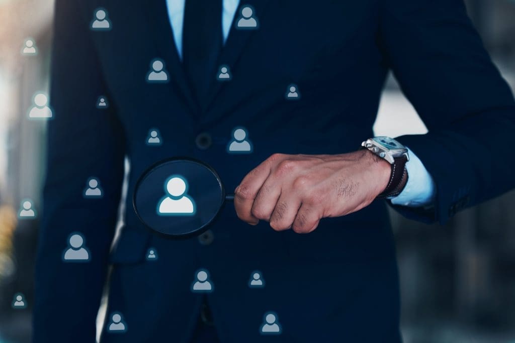 A person in a suit holding a magnifying glass, focusing on a digital icon of a human figure among various similar icons. The scene symbolizes the concept of searching, recruitment, or selection in a professional context, akin to using tools like ZoomInfo for precise candidate identification while respecting opt-out preferences.