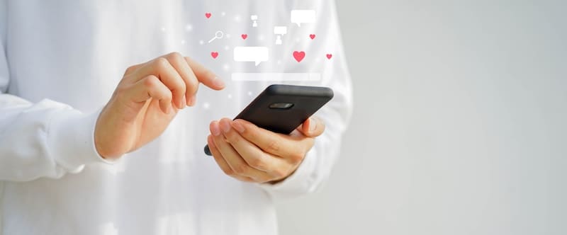 Person in a white shirt holding a smartphone with floating icons of hearts, chat bubbles, and search symbols above the screen, suggesting social media interaction and online dating tips. Blurred gray background.