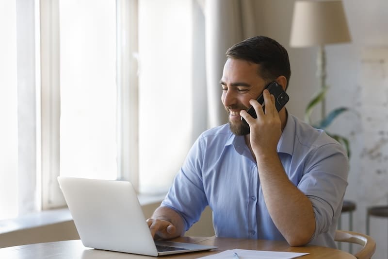 Happy busy professional man talking on telephone, making call to client, giving consultation, typing on laptop, using online application at workplace, negotiating on business project on mobile phone.