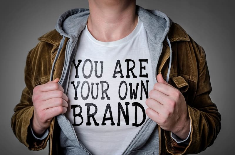 A person opens their brown jacket and gray hoodie to reveal a white t-shirt with "YOU ARE YOUR OWN BRAND" in bold black letters, highlighting the importance of self-image—much like google reputation management for individuals.