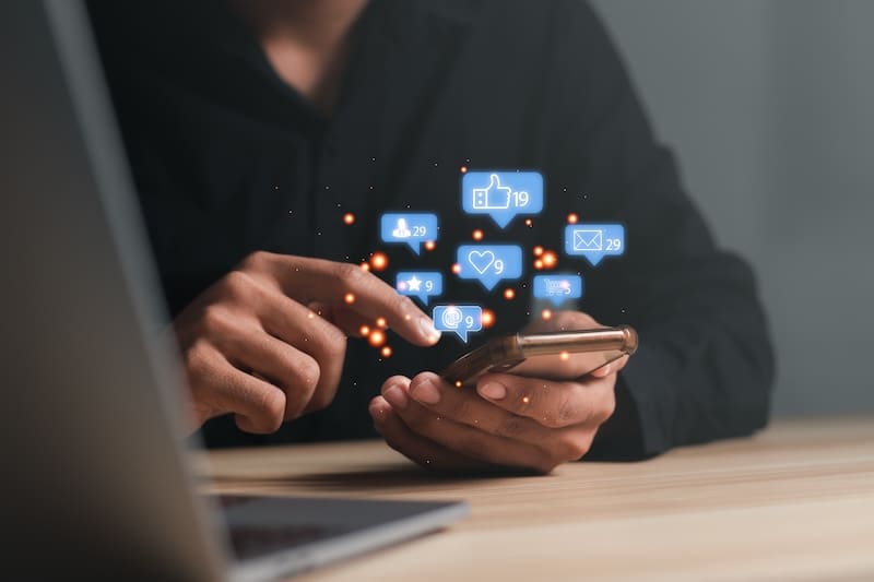 A person wearing a dark shirt is using a smartphone at a wooden desk, focusing on reputation management for lawyers. Various social media notification icons, such as thumbs up, hearts, and message bubbles, are floating around the phone, indicating engagement with social media content.