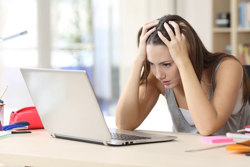 A woman sits at a desk, hands on her head, looking frustrated at a laptop. The cluttered desk, with stationery and a red pencil case, echoes her stress as she focuses intensely on suppressing Google search results displayed on the screen.